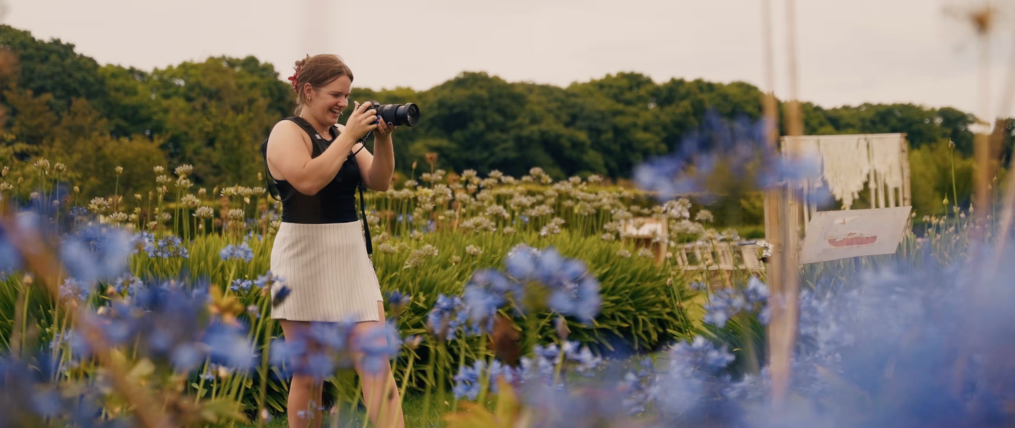 Huwelijksfotograaf Ines staat tussen de paarse bloemen en kijkt al lachend door haar lens.